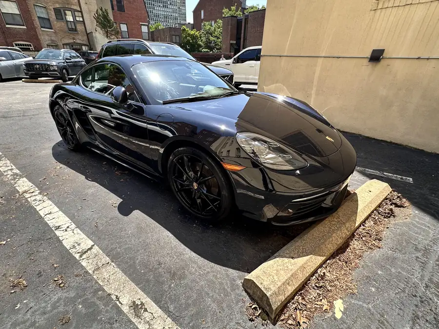 Black Porsche Cayman parked in a city lot, its paint wiped to a gloss in full sun